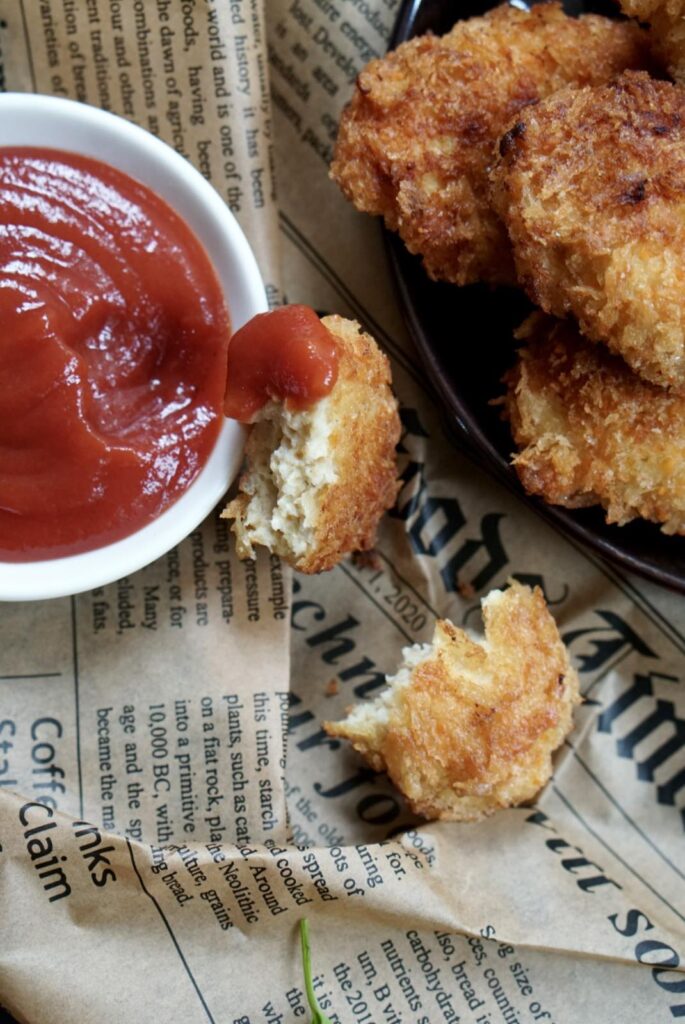 A broken piece of nugget dipped in ketchup beside a pile of crispy nuggets.