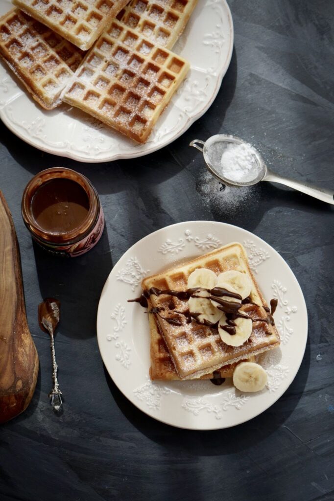 A top-down view of a breakfast table setting on a dark surface. In the foreground, a white plate holds a crispy homemade waffle topped with banana slices, chocolate drizzle, and powdered sugar. In the background, there is a plate with two plain waffles, a small pot of chocolate spread on a rustic wooden board, and a powdered sugar strainer.