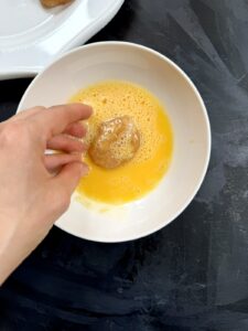 A hand dipping a shaped chicken nugget into a bowl of beaten egg.