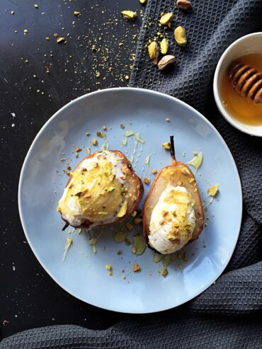 Two halves of a baked pear topped with melting vanilla ice cream, crushed pistachios, and honey on a light blue plate.