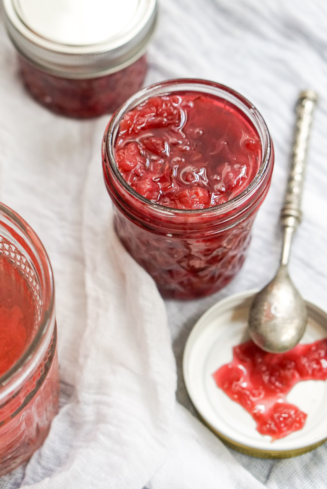 A glass jar filled with bright pink easy homemade rose petal jam next to a silver spoon and a small plate on a white cloth