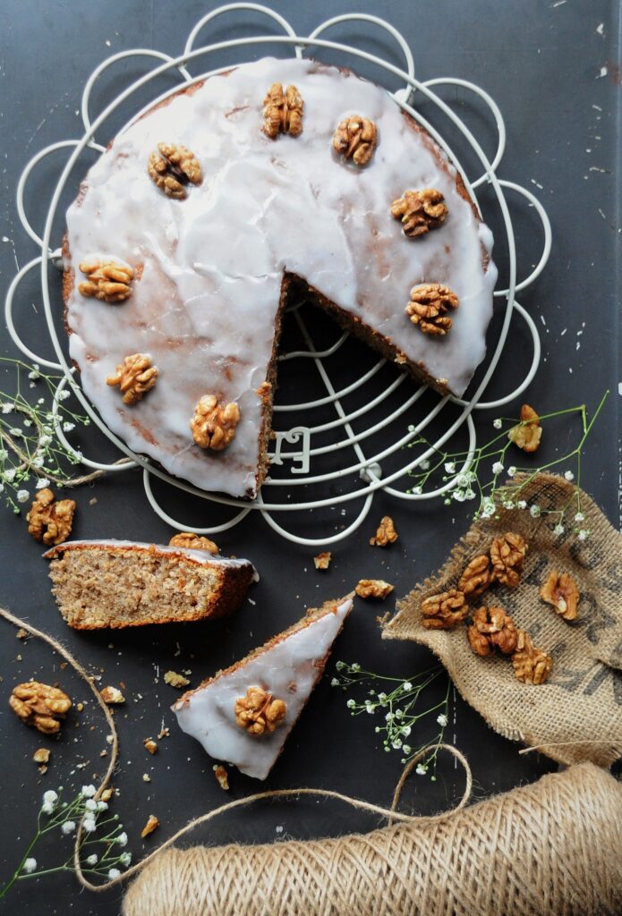 Top-down view of a sliced sour cream walnut cake covered in white vanilla glaze and walnut halves, placed on a wire cooling rack with a cut slice resting in the foreground.
