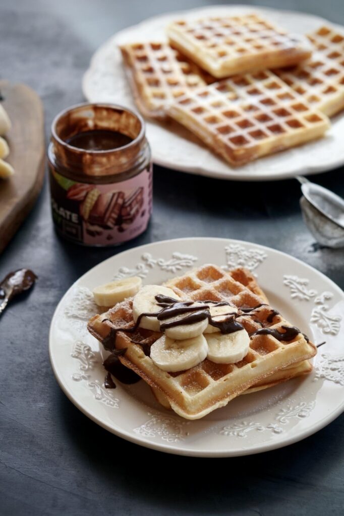 A close-up of an easy homemade waffle topped with fresh banana slices and chocolate hazelnut spread on a white decorative plate, with a jar of chocolate spread in the background.