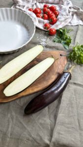 Raw eggplant halves in a white dish with fresh cherry tomatoes, mozzarella cheese packets, and a wooden cutting board on a linen cloth, representing ingredients for baked eggplant.