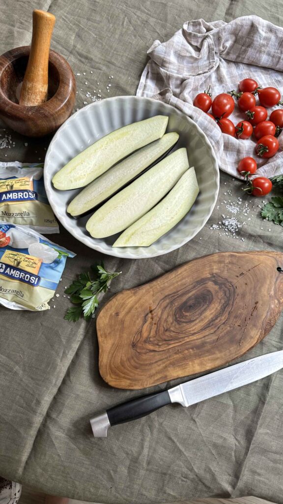 Raw eggplant halves in a white dish with fresh cherry tomatoes, mozzarella cheese packets, and a wooden cutting board on a linen cloth, representing ingredients for baked eggplant.