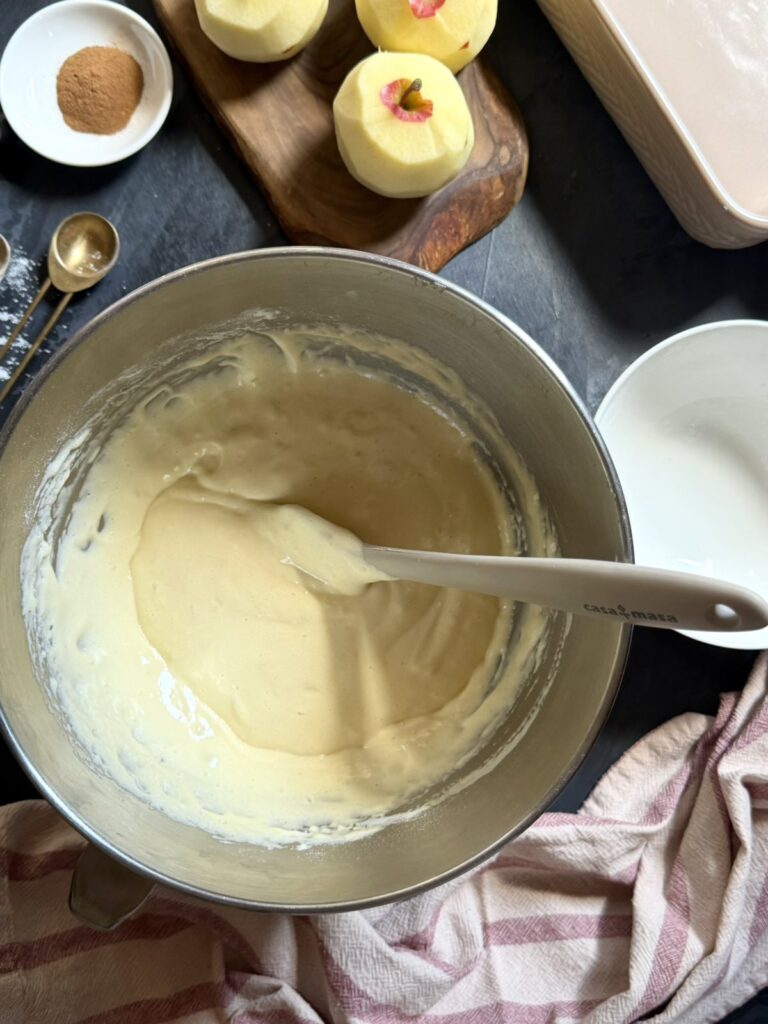 Fluffy cake batter in a mixing bowl with peeled apples nearby