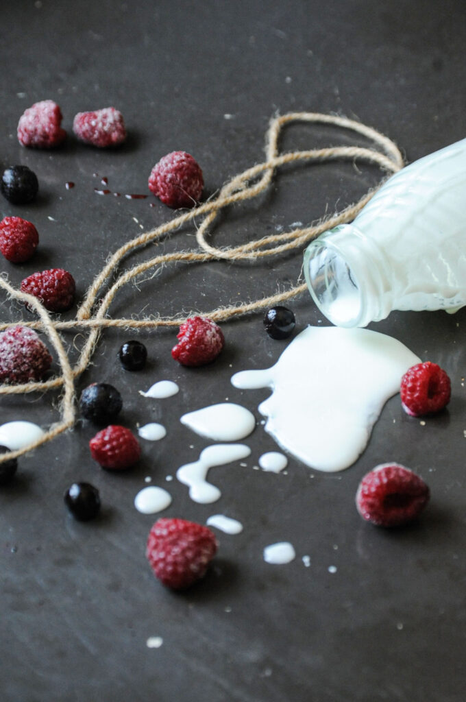 A small glass bottle tipped over, spilling white yogurt onto a dark rustic surface, surrounded by scattered raspberries, black currants, and rustic twine.