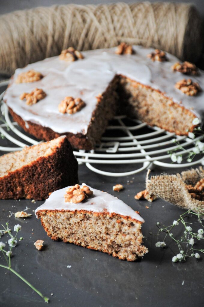A slice of moist sour cream walnut cake topped with sweet vanilla glaze and walnut halves, sitting next to the whole cake on a wire cooling rack.