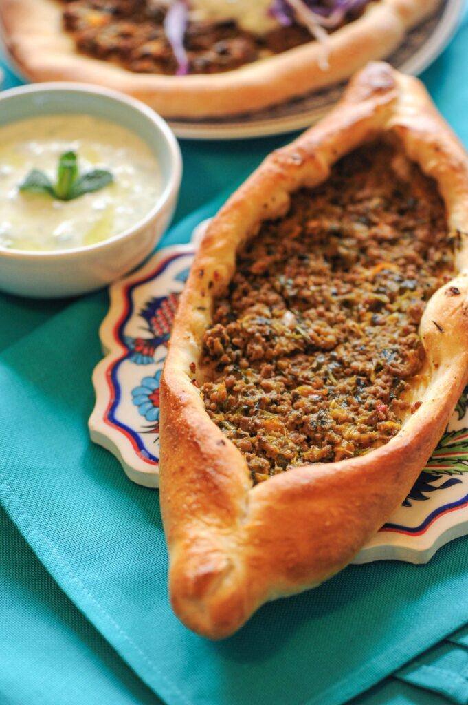 A close-up of a baked Turkish pide flatbread filled with seasoned ground beef, served on a patterned plate next to a bowl of yogurt sauce.