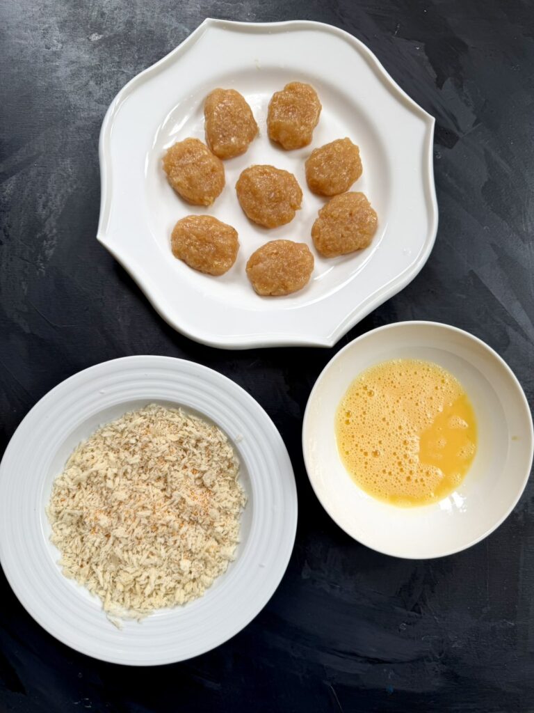 Shaped raw ground chicken nuggets on a plate next to a bowl of beaten egg and a plate of breadcrumbs.