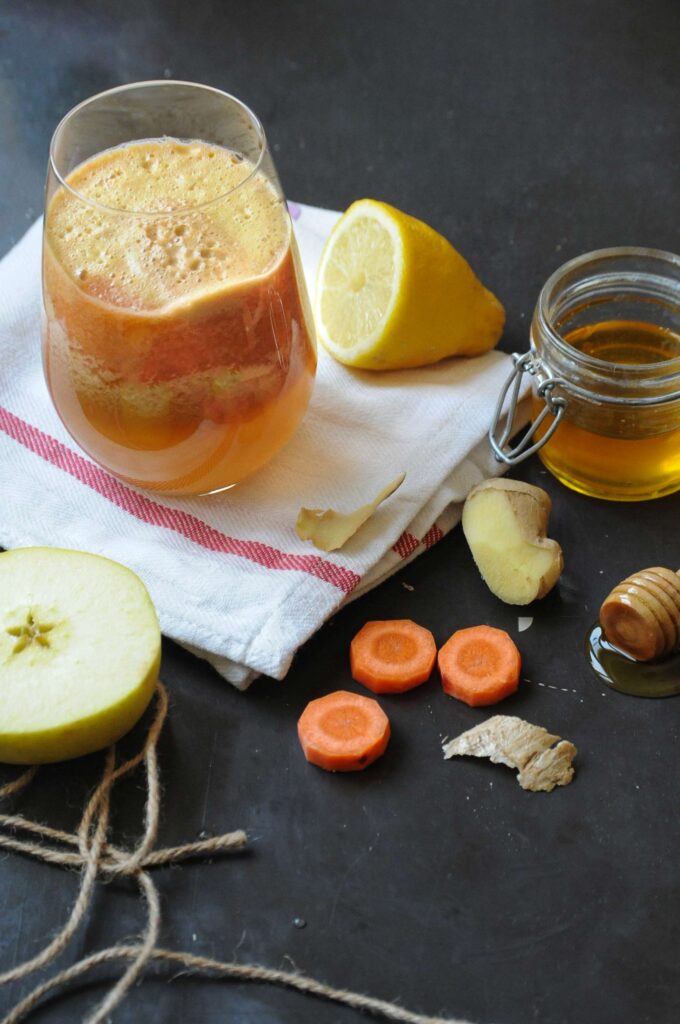 A tall glass of fresh homemade carrot apple ginger juice on a white napkin, surrounded by a halved green apple, sliced carrots, ginger root, a lemon half, and a jar of honey with a wooden dipper on a dark surface.