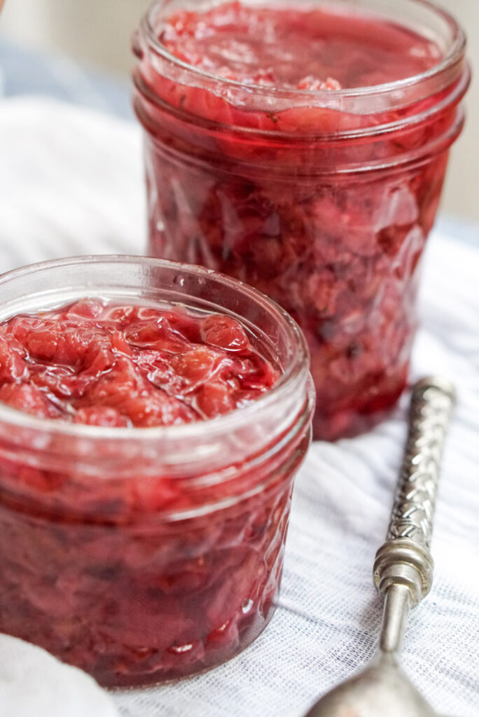 Two glass jars filled with bright pink homemade rose petal jam on a white cloth next to a vintage silver spoon
