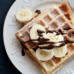 Top-down view of a square homemade waffle on a white decorative plate, topped with fresh banana slices, chocolate drizzle, and a dusting of powdered sugar on a dark background.