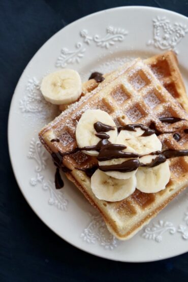 Top-down view of a square homemade waffle on a white decorative plate, topped with fresh banana slices, chocolate drizzle, and a dusting of powdered sugar on a dark background.
