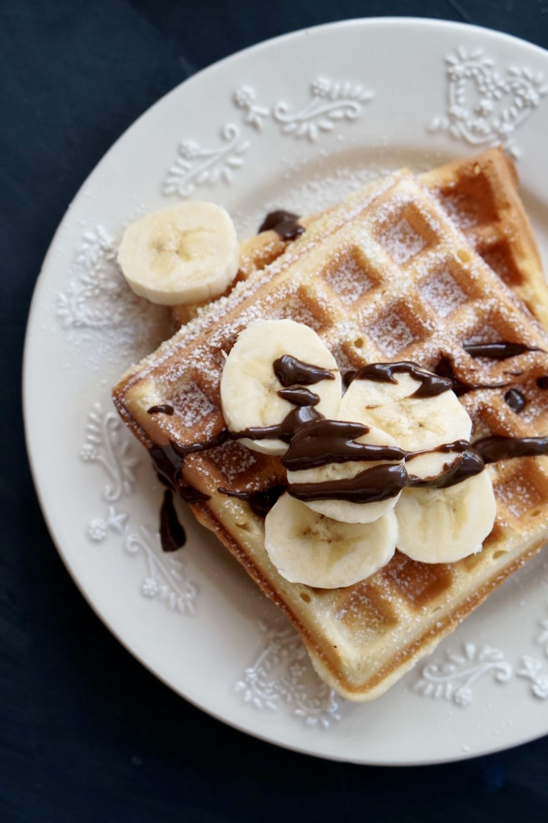 Top-down view of a square homemade waffle on a white decorative plate, topped with fresh banana slices, chocolate drizzle, and a dusting of powdered sugar on a dark background.