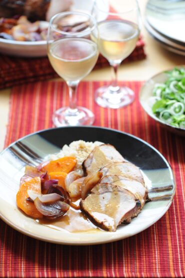 Plate of sliced pork, roasted squash, and red onion on a dinner table with glasses of white wine