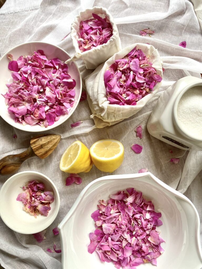 Fresh pink rose petals in white bowls, halved lemons, and a wooden citrus reamer on a white cloth for making rose petal jam