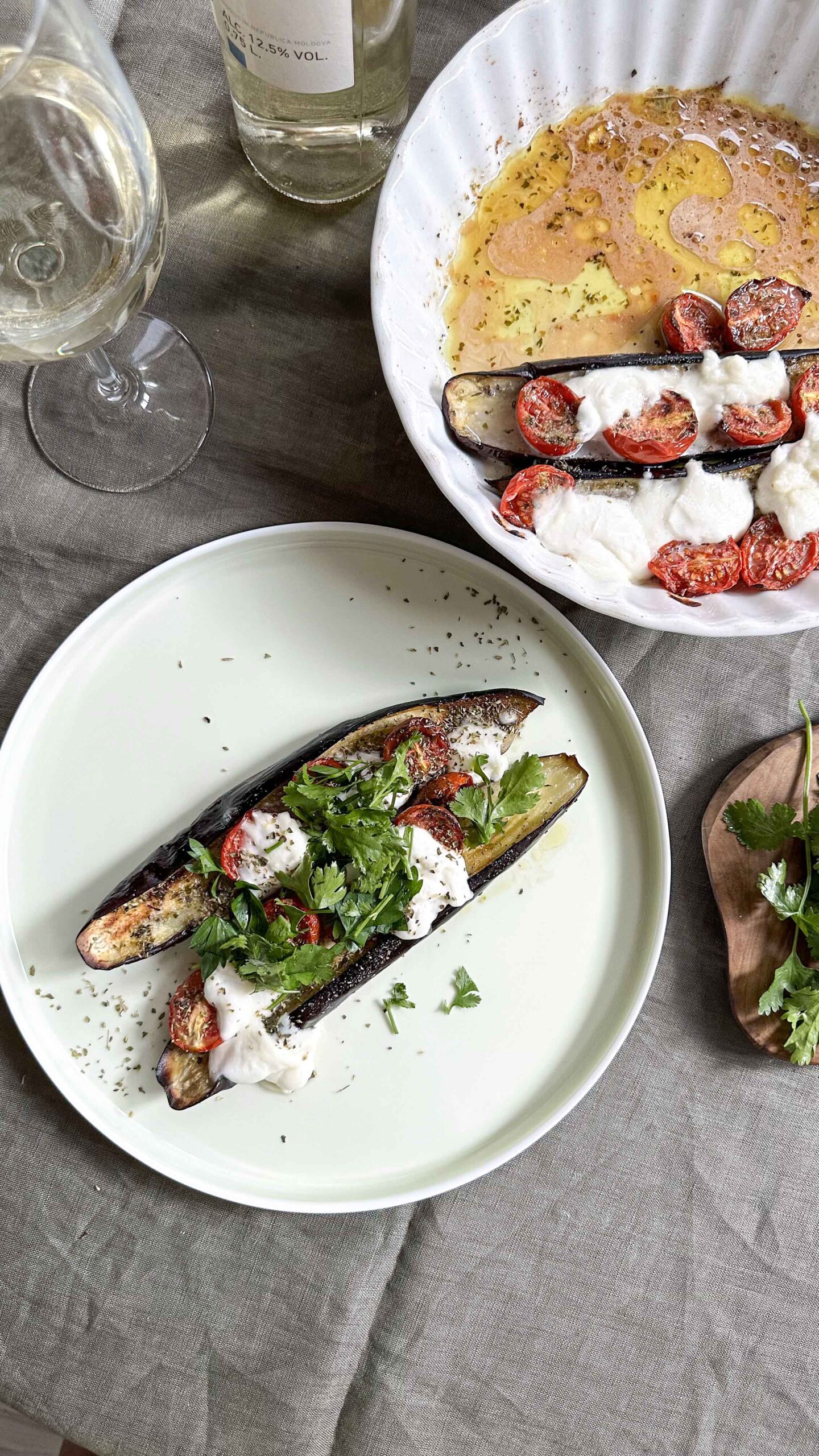 Top-down view of two white plates serving roasted eggplant halves topped with mozzarella, tomatoes, and fresh herbs, arranged on a grey linen tablecloth alongside a glass of white wine.