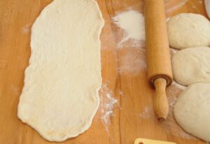 An oval piece of rolled-out dough next to a wooden rolling pin and three round dough balls on a flour-dusted wooden surface