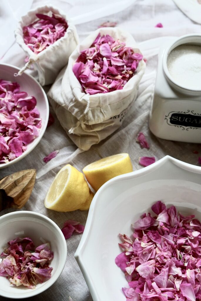 Flat lay of pink rose petals, fresh lemon halves, and a vintage sugar bowl on a linen cloth for making rose petal jam