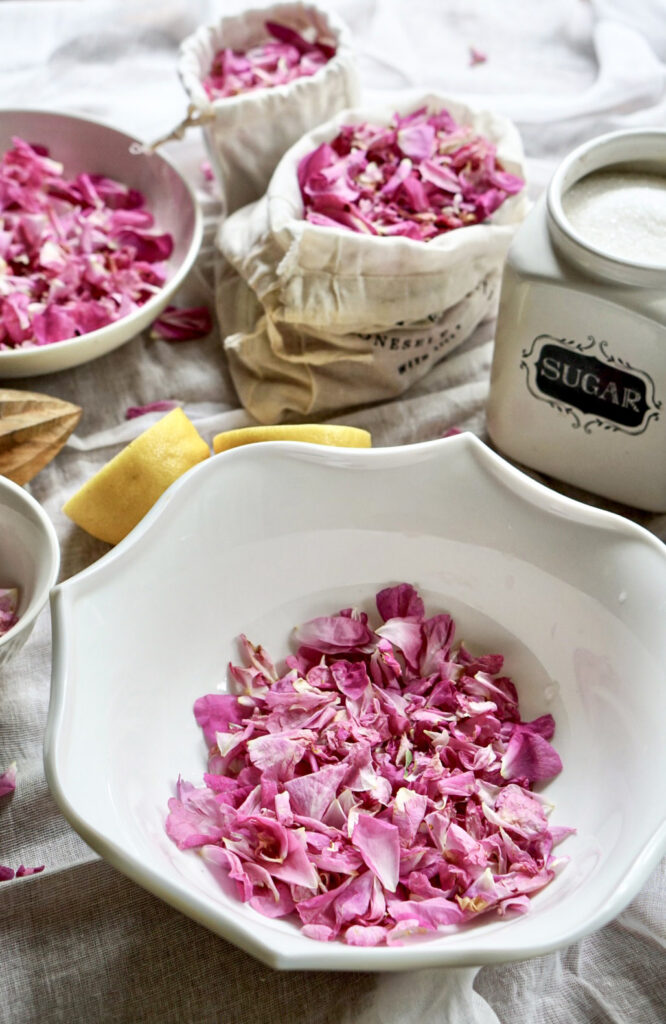 Fresh pink rose petals in a white bowl, lemon wedges, and a vintage sugar container on a white cloth for making rose petal jam
