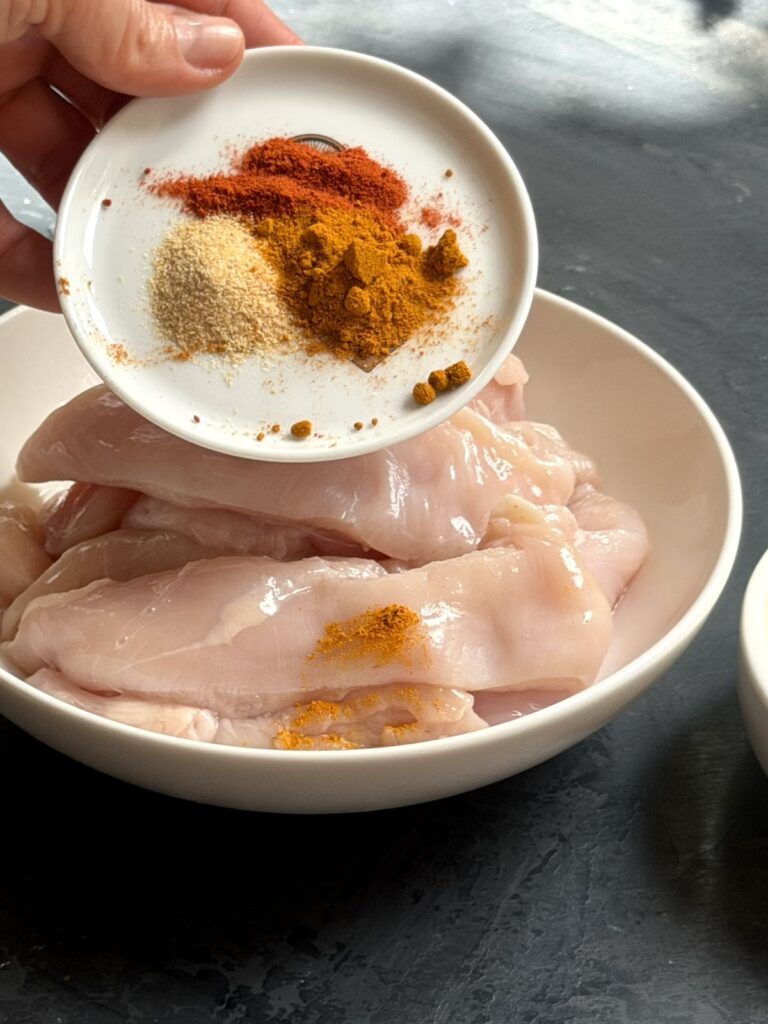 Adding spices to sliced chicken breast before pan-searing.