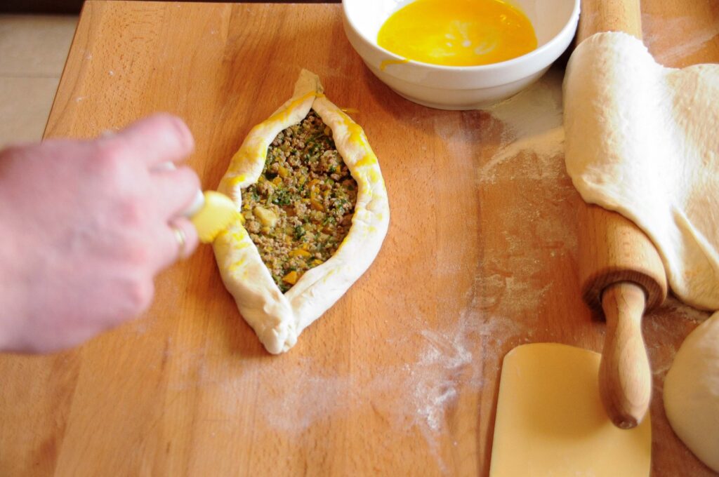 A hand using a pastry brush to apply egg wash to the folded edges of a raw Turkish pide stuffed with ground beef on a wooden board