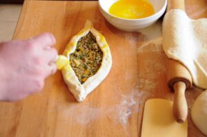 A hand using a pastry brush to apply egg wash to the folded edges of a raw Turkish pide stuffed with ground beef on a wooden board