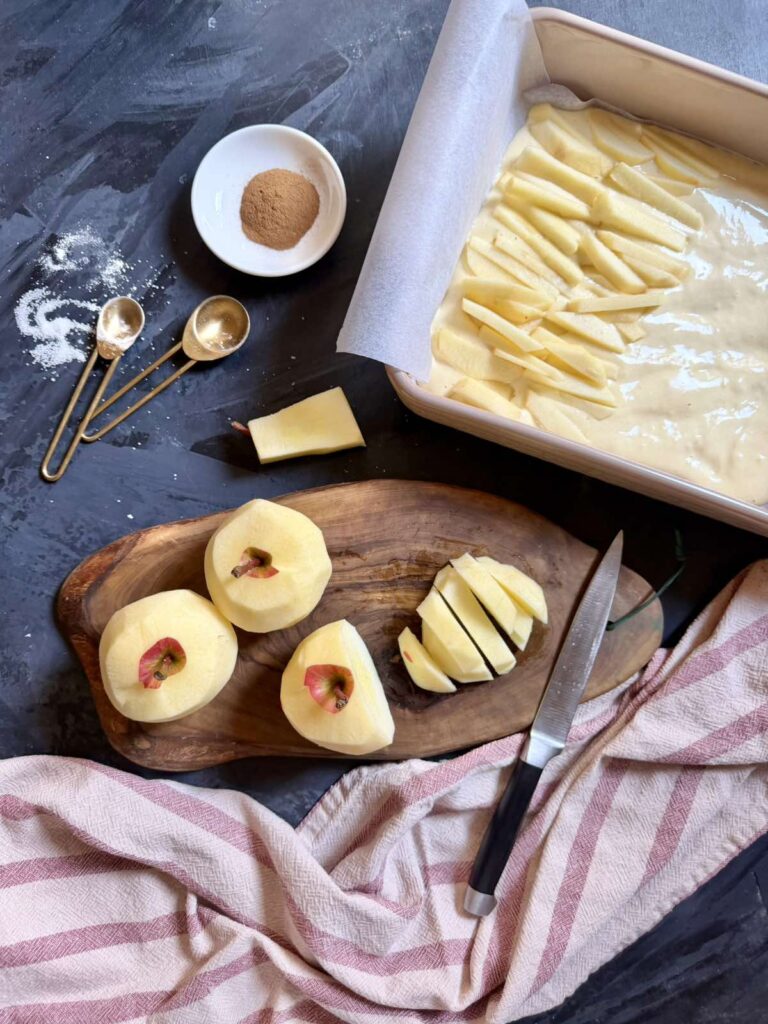 Thinly sliced apples on a wooden board beside a pan of cake batter