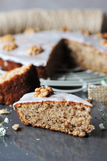 A close-up front view of a slice of sour cream walnut cake topped with vanilla glaze and a walnut, resting on a dark surface with the rest of the cake softly blurred in the background.