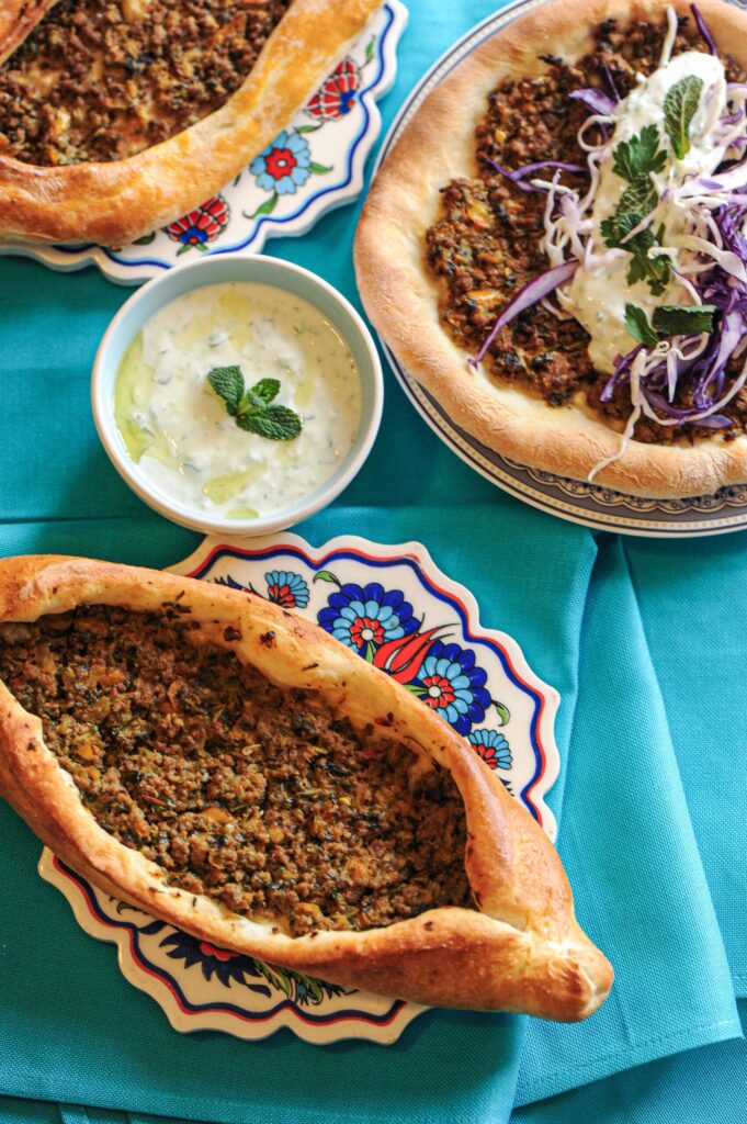 Three Turkish pides with ground beef and vegetable filling on patterned plates, served with yogurt sauce and red cabbage salad over a bright blue tablecloth