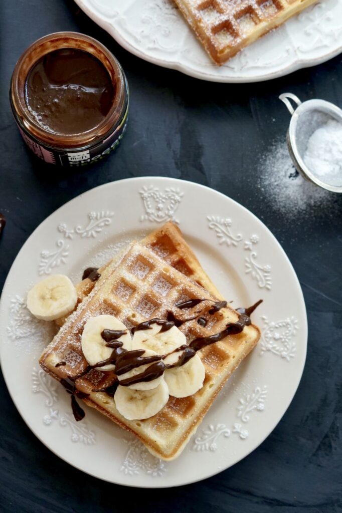 A top-down view of a square homemade waffle topped with fresh banana slices and chocolate drizzle on a white patterned plate, with a jar of chocolate spread and powdered sugar in the dark background.