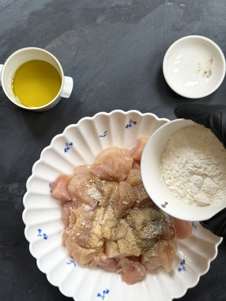 Adding cornstarch to seasoned raw chicken pieces in a bowl.