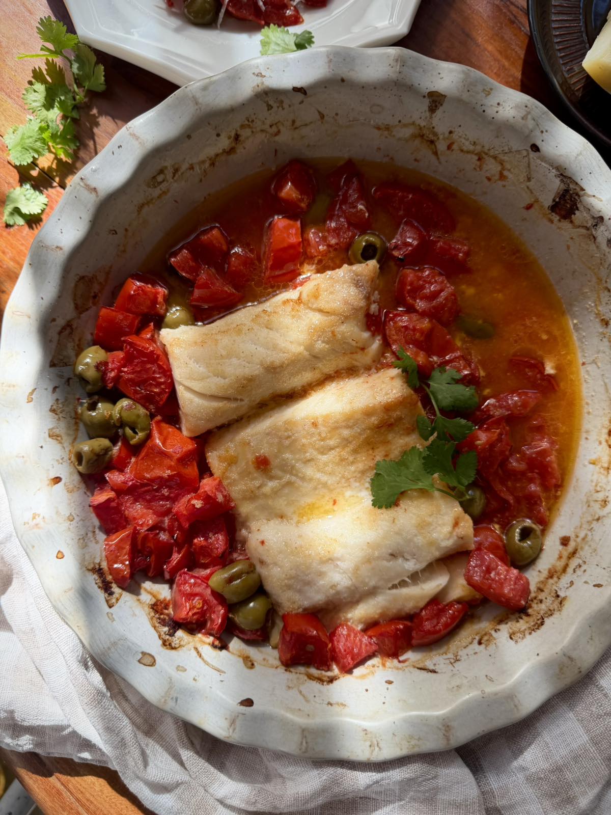Close-up of baked cod in tomato juices with green olives and fresh herb leaves