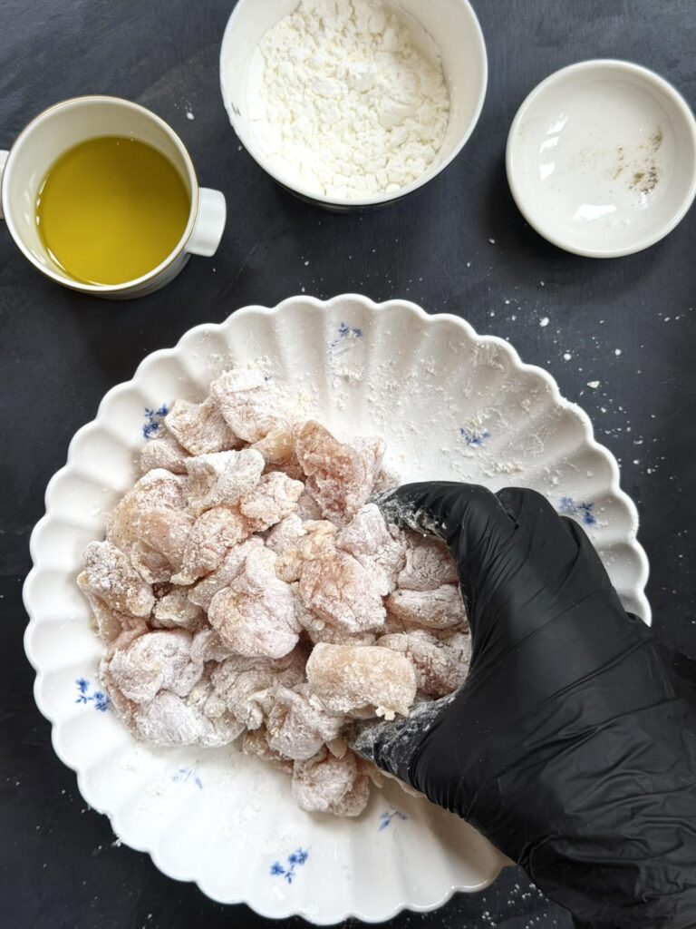 A gloved hand mixing chicken pieces with cornstarch in a bowl.