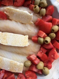Close-up of raw cod fillets with butter slices, chopped tomatoes, and green olives
