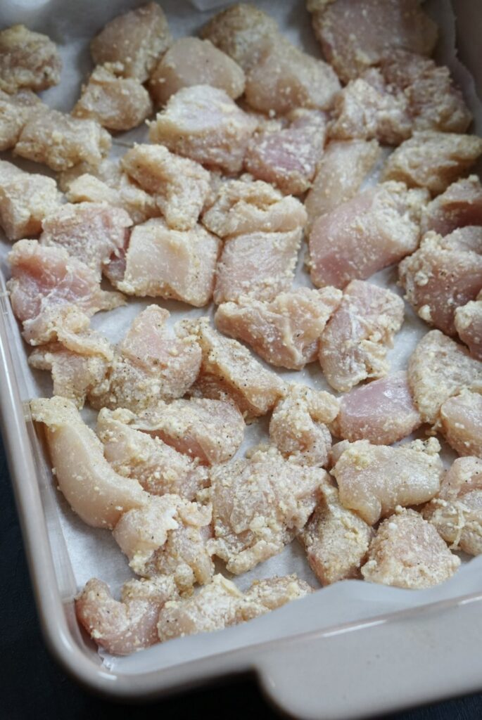 Cornstarch-coated raw chicken pieces arranged in a parchment-lined baking dish.