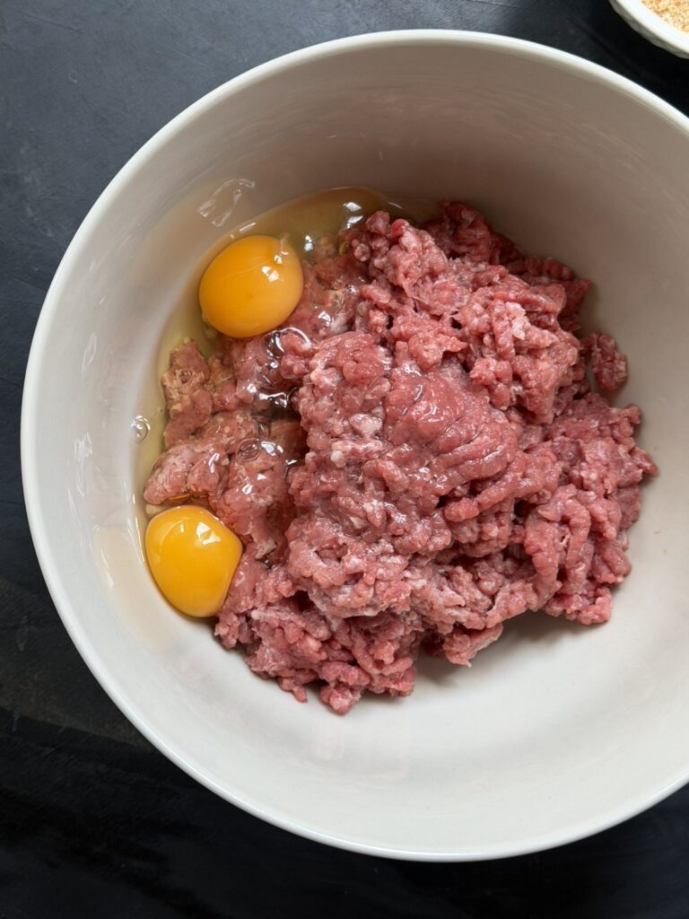 Ground beef with two cracked eggs in a white mixing bowl before mixing