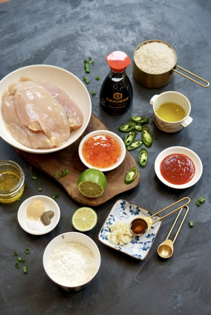 Ingredients for Korean-inspired baked chicken, including raw chicken, soy sauce, cornstarch, oil, honey, chili sauces, lime, green chilies, and seasonings.