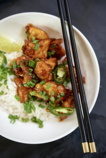 Sweet and spicy chicken over white rice in a bowl, topped with green onions and sliced green chili, with a lime wedge and chopsticks.