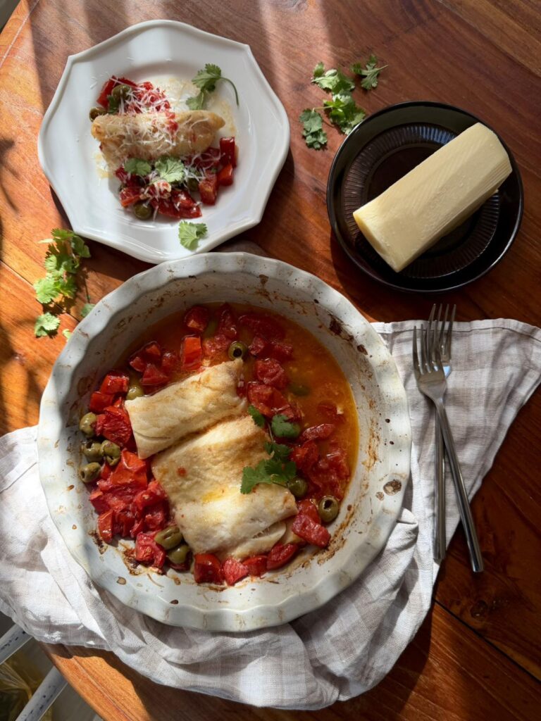 Oven-baked cod with tomatoes and olives in a baking dish, with a plated serving and a block of Parmesan