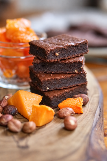 A stack of chocolate pumpkin hazelnut brownies on a wooden board with pumpkin pieces and hazelnuts