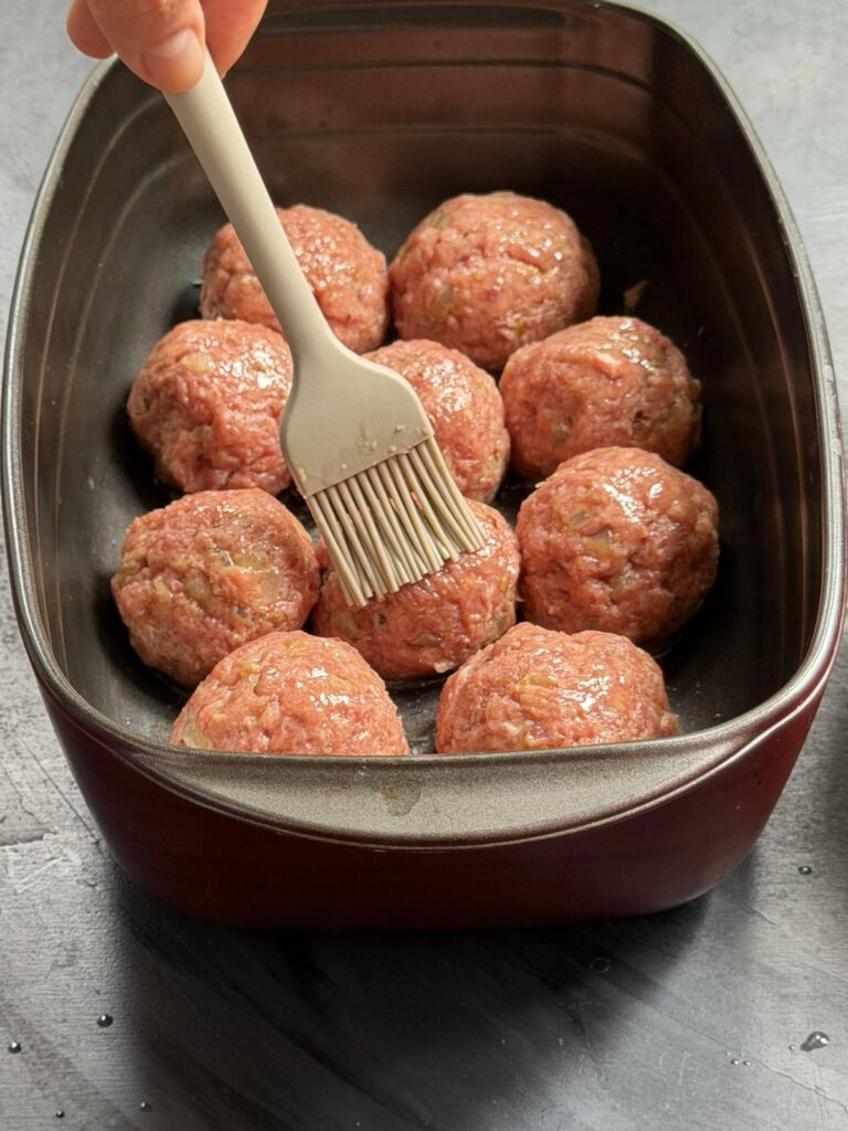 Brushing raw beef patties in a baking dish with a pastry brush before baking
