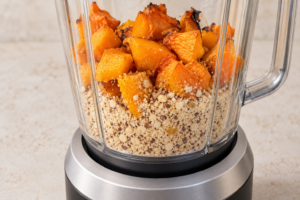 Roasted orange pumpkin chunks on top of ground hazelnuts inside a clear blender jar