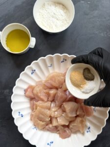 A gloved hand adding salt, black pepper, and garlic powder to raw chicken pieces in a bowl.