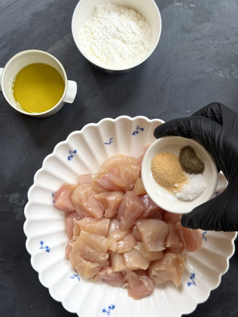 A gloved hand adding salt, black pepper, and garlic powder to raw chicken pieces in a bowl.