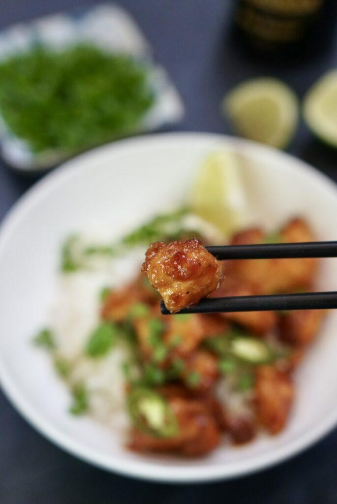 Chopsticks holding a glazed chicken bite above a bowl of chicken and rice.